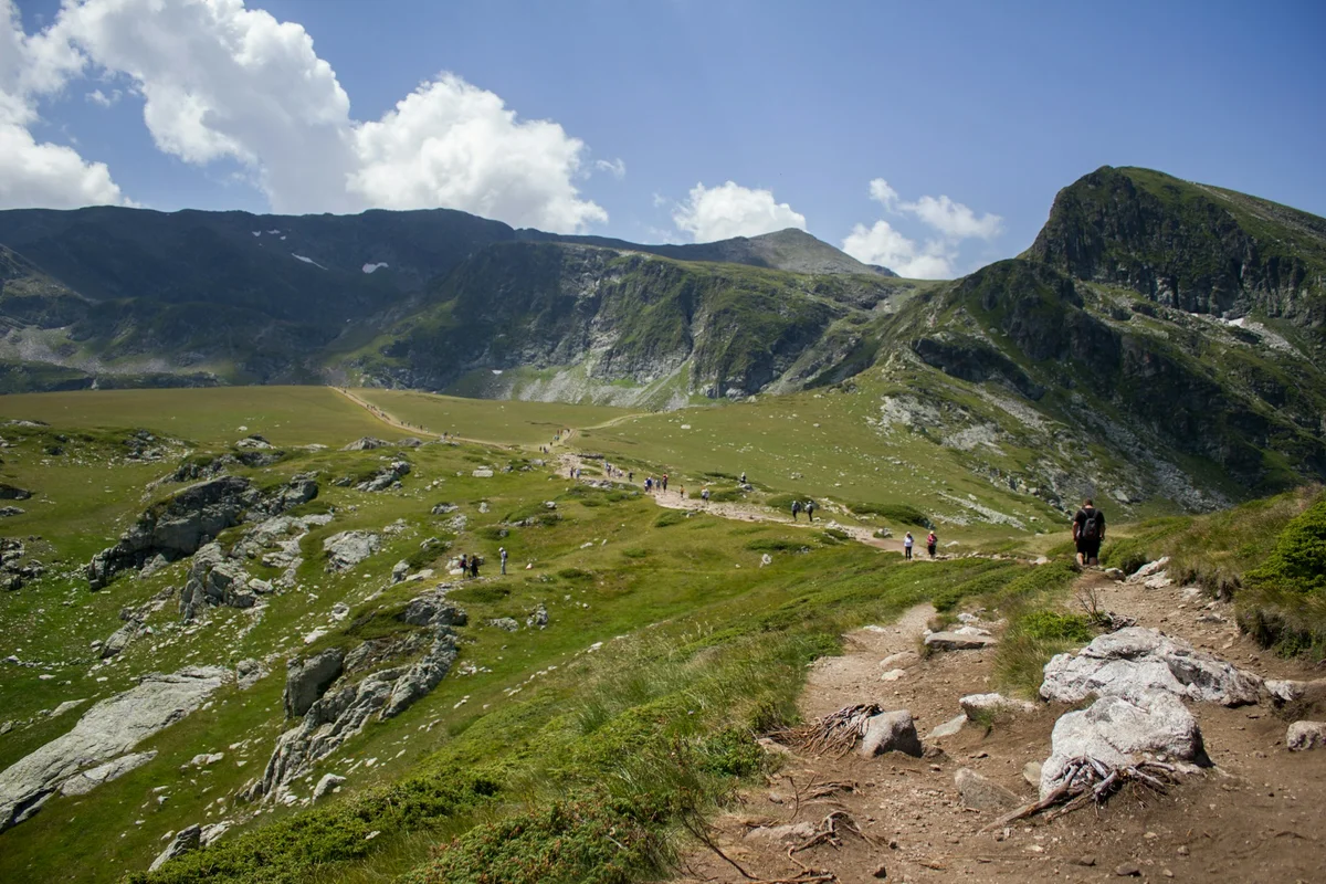 Hikers walking along a winding mountain trail through green alpine hills toward the rocky peaks of the Rila Mountains