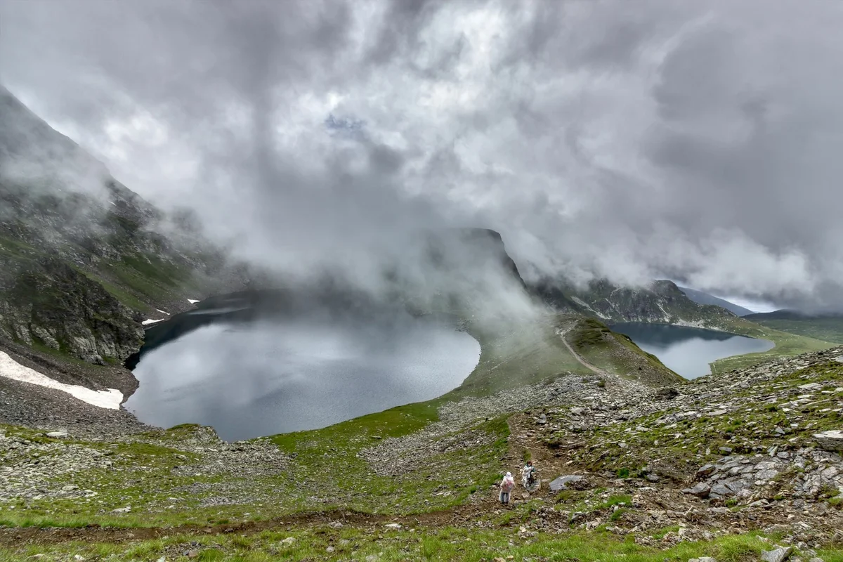 Clouds and mist swirling over glacial lakes and rocky slopes in the Seven Rila Lakes, with hikers on a mountain trail